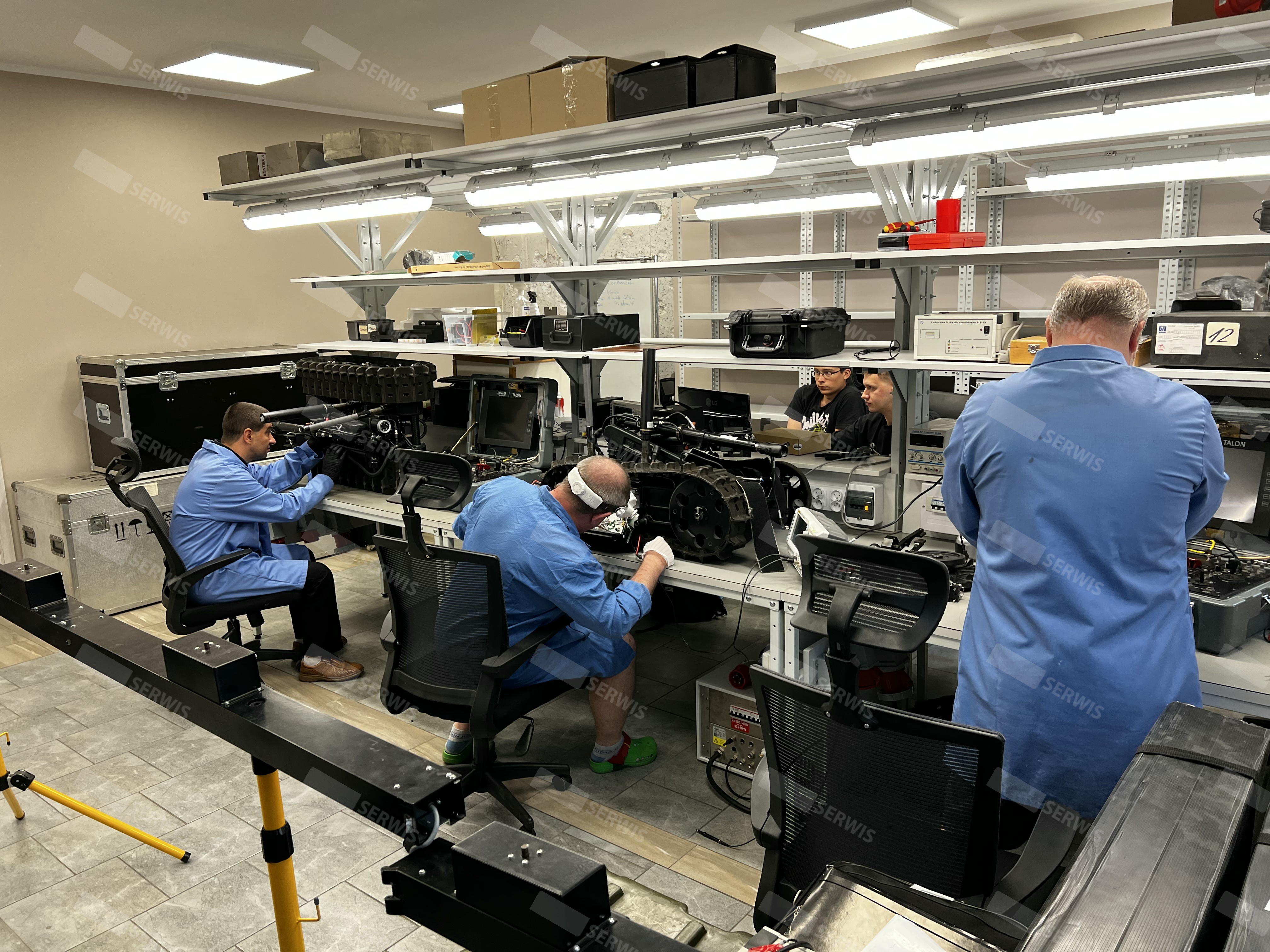 Technicy w niebieskich fartuchach naprawiają sprzęt elektroniczny w warsztacie serwisowym