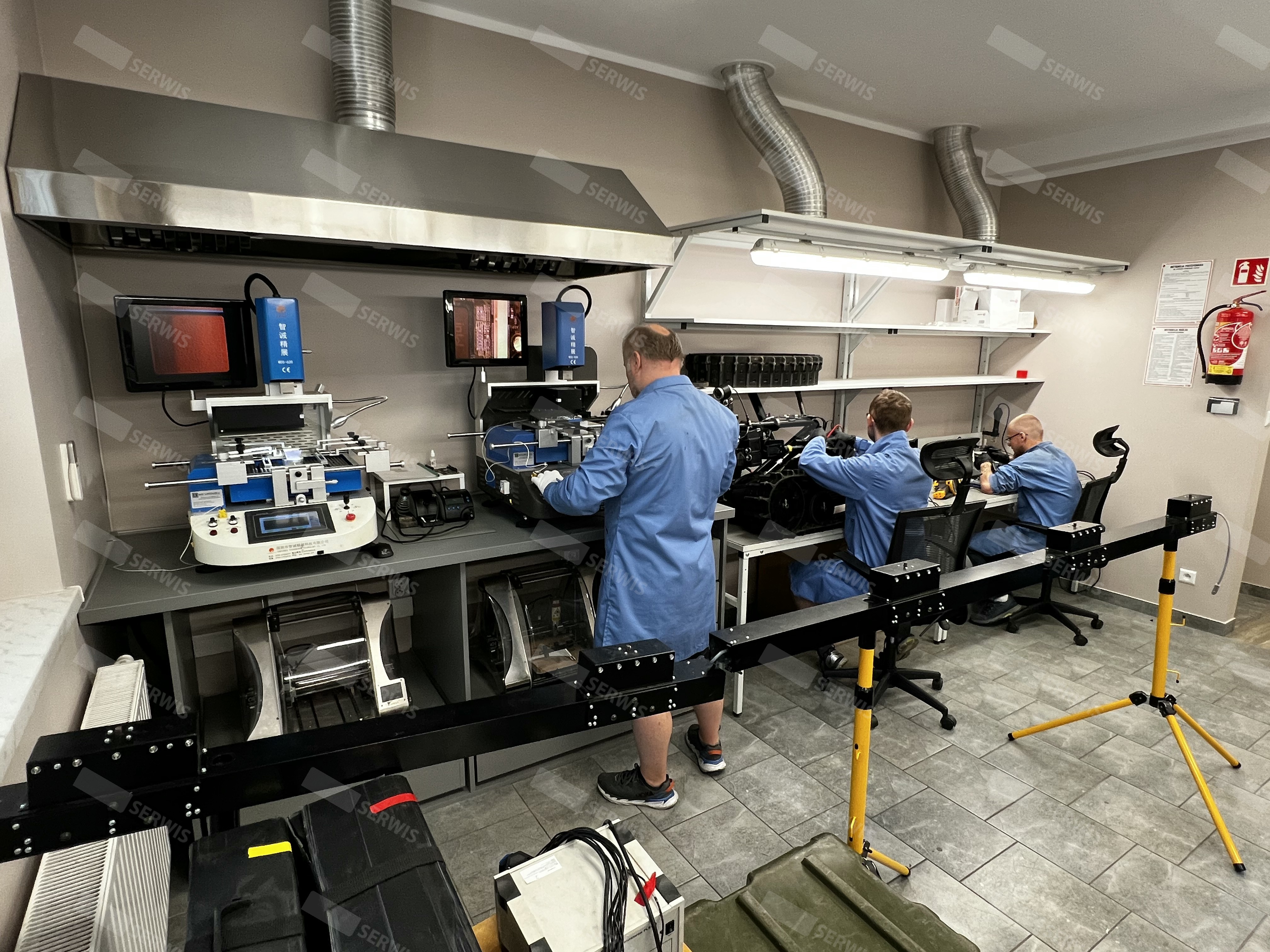 Technicy w niebieskich fartuchach naprawiają sprzęt elektroniczny w laboratorium serwisowym